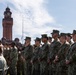 A large formation of U.S. Navy personnel in NWU Type III (woodland digital camouflage) uniforms stand at attention during what appears to be an outdoor military ceremony or formation. In the background, a prominent red-brick clock tower and a flagpole with a flag at half-staff are visible. Civilian observers are seated to the left, suggesting this is a formal commemorative or official event.