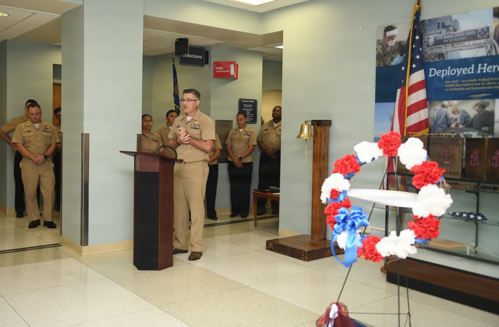 A military officer in tan/khaki uniform stands at a wooden podium delivering a speech during what appears to be a memorial or commemorative ceremony. Several uniformed Navy personnel stand at attention in the background near a 'Collections' office area. The foreground features a red, white, and blue floral wreath, an American flag, a brass bell on a pedestal, and a 'Deployed Heroes' display banner.