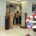 A military officer in tan/khaki uniform stands at a wooden podium delivering a speech during what appears to be a memorial or commemorative ceremony. Several uniformed Navy personnel stand at attention in the background near a 'Collections' office area. The foreground features a red, white, and blue floral wreath, an American flag, a brass bell on a pedestal, and a 'Deployed Heroes' display banner.