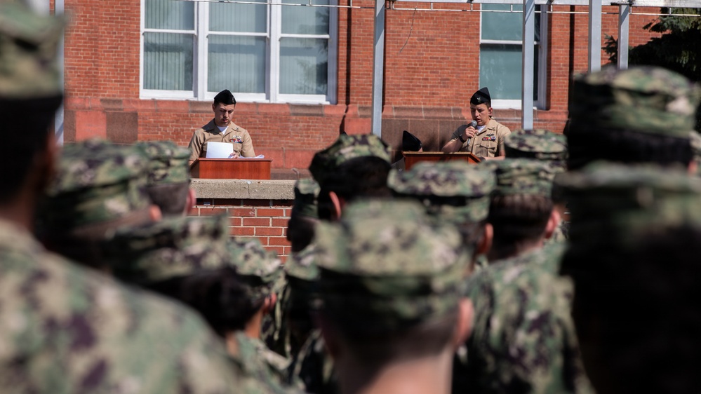 A military formation or ceremony is taking place outdoors in front of a red brick building. Two individuals in tan/khaki uniforms stand at separate podiums, appearing to address a large group of personnel in camouflage uniforms. The scene suggests an official military event such as a change of command, graduation, or formal briefing.