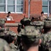 A military formation or ceremony is taking place outdoors in front of a red brick building. Two individuals in tan/khaki uniforms stand at separate podiums, appearing to address a large group of personnel in camouflage uniforms. The scene suggests an official military event such as a change of command, graduation, or formal briefing.