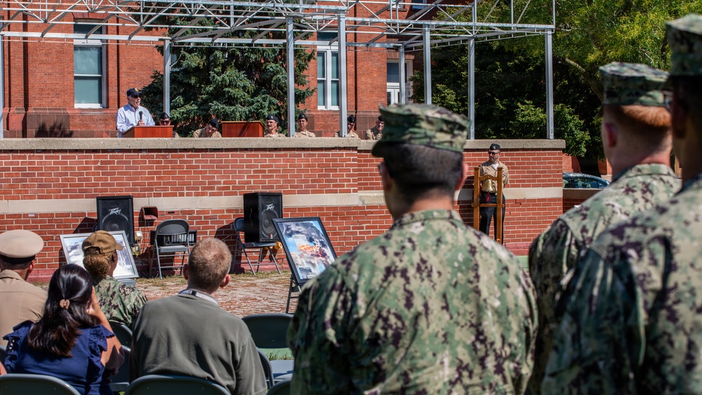 An outdoor military ceremony is taking place at what appears to be a naval installation, with a civilian speaker addressing an audience from an elevated podium on a brick stage. Personnel in US Navy blue digital camouflage (NWU Type III) uniforms stand in formation while seated attendees, including civilians and military members, observe the proceedings. Framed photographs or memorial displays are visible near the stage area, suggesting a commemorative or memorial event.