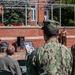 An outdoor military ceremony is taking place at what appears to be a naval installation, with a civilian speaker addressing an audience from an elevated podium on a brick stage. Personnel in US Navy blue digital camouflage (NWU Type III) uniforms stand in formation while seated attendees, including civilians and military members, observe the proceedings. Framed photographs or memorial displays are visible near the stage area, suggesting a commemorative or memorial event.