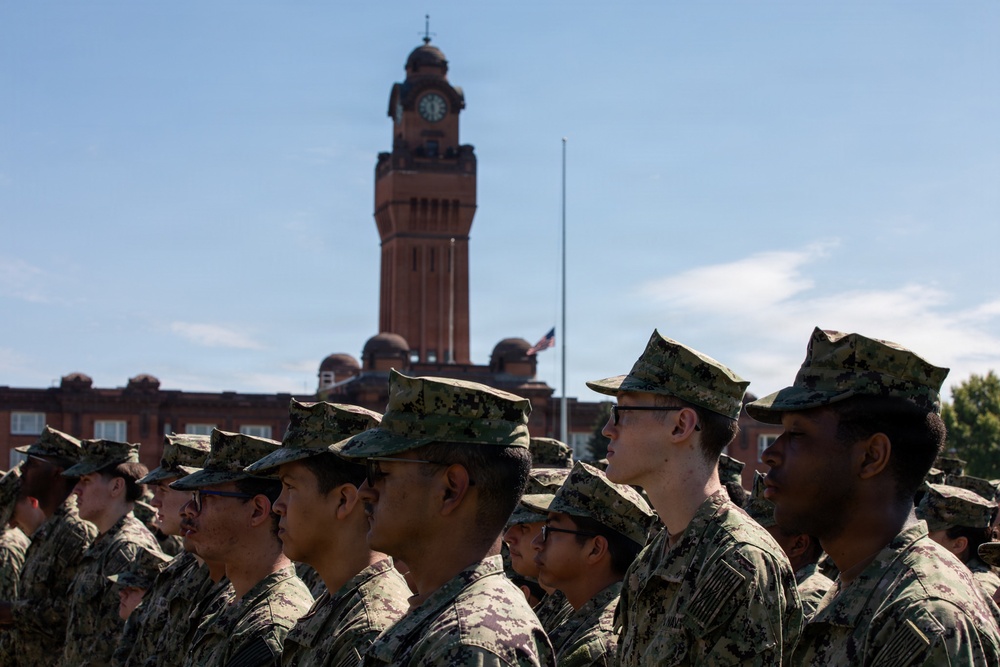A formation of military personnel in digital camouflage uniforms stands at attention on a parade ground. Behind them stands a prominent red brick clock tower building, consistent with a military installation or academy. An American flag is visible at half-staff on a flagpole in the background.