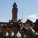 A formation of military personnel in digital camouflage uniforms stands at attention on a parade ground. Behind them stands a prominent red brick clock tower building, consistent with a military installation or academy. An American flag is visible at half-staff on a flagpole in the background.