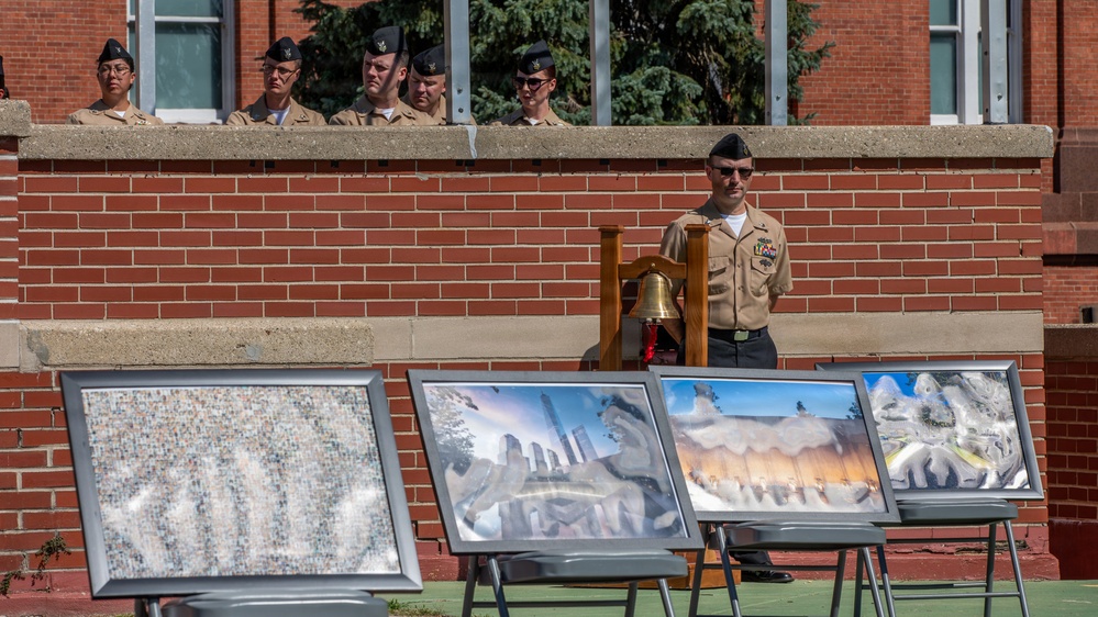 A military ceremony is taking place outdoors against a red brick building, with a uniformed service member standing at attention beside a ceremonial bell on a wooden stand. Several other military personnel in tan/khaki uniforms and black garrison caps observe from an elevated position above a concrete ledge. In the foreground, multiple framed photographs or artwork panels are displayed on easels, appearing to depict 9/11-related imagery including the Twin Towers and memorials.