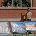 A military ceremony is taking place outdoors against a red brick building, with a uniformed service member standing at attention beside a ceremonial bell on a wooden stand. Several other military personnel in tan/khaki uniforms and black garrison caps observe from an elevated position above a concrete ledge. In the foreground, multiple framed photographs or artwork panels are displayed on easels, appearing to depict 9/11-related imagery including the Twin Towers and memorials.