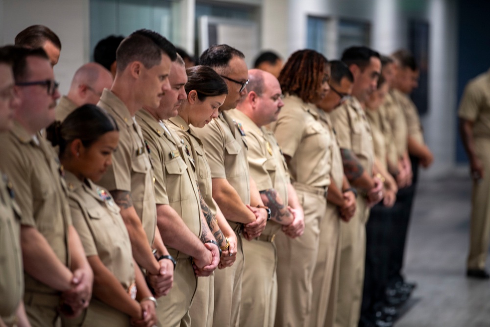 A group of military personnel in matching tan/khaki uniforms are standing in formation with heads bowed and hands clasped in front of them, suggesting a moment of silence or prayer during a formal ceremony. The group is diverse in gender and ethnicity, and appears to be indoors in a modern facility. The solemn posture and uniformity suggest a commemorative or official military event.