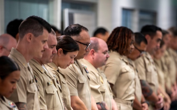 A group of military personnel in matching tan/khaki uniforms are standing in formation with heads bowed and hands clasped in front of them, suggesting a moment of silence or prayer during a formal ceremony. The group is diverse in gender and ethnicity, and appears to be indoors in a modern facility. The solemn posture and uniformity suggest a commemorative or official military event.
