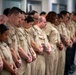 A group of military personnel in matching tan/khaki uniforms are standing in formation with heads bowed and hands clasped in front of them, suggesting a moment of silence or prayer during a formal ceremony. The group is diverse in gender and ethnicity, and appears to be indoors in a modern facility. The solemn posture and uniformity suggest a commemorative or official military event.