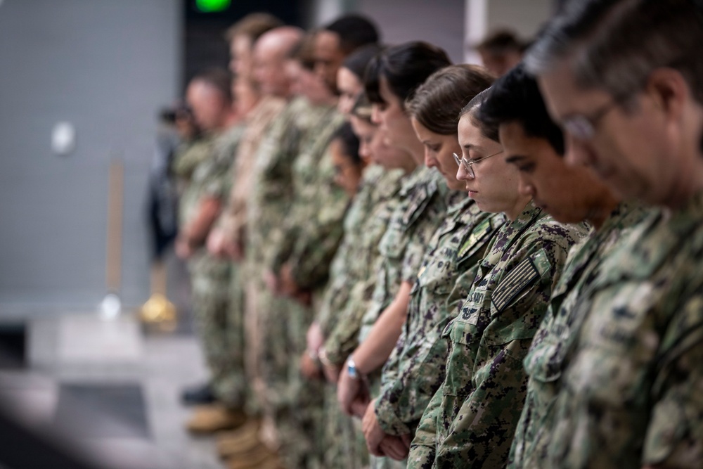 A group of U.S. Navy personnel in NWU Type III (woodland digital camouflage) uniforms stand in a line with heads bowed, suggesting a moment of prayer, silence, or invocation during a formal ceremony. The individuals are arranged in a single file facing left, with the foreground subjects in sharp focus and background figures progressively blurred. A ceremonial object, possibly a bell or trophy, is partially visible on a surface to the left.