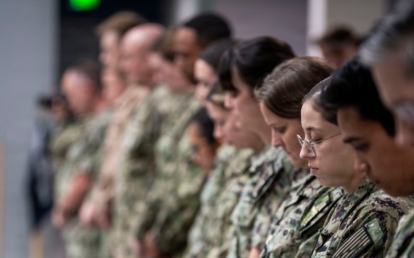 A group of U.S. Navy personnel in NWU Type III (woodland digital camouflage) uniforms stand in a line with heads bowed, suggesting a moment of prayer, silence, or invocation during a formal ceremony. The individuals are arranged in a single file facing left, with the foreground subjects in sharp focus and background figures progressively blurred. A ceremonial object, possibly a bell or trophy, is partially visible on a surface to the left.