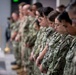 A group of U.S. Navy personnel in NWU Type III (woodland digital camouflage) uniforms stand in a line with heads bowed, suggesting a moment of prayer, silence, or invocation during a formal ceremony. The individuals are arranged in a single file facing left, with the foreground subjects in sharp focus and background figures progressively blurred. A ceremonial object, possibly a bell or trophy, is partially visible on a surface to the left.