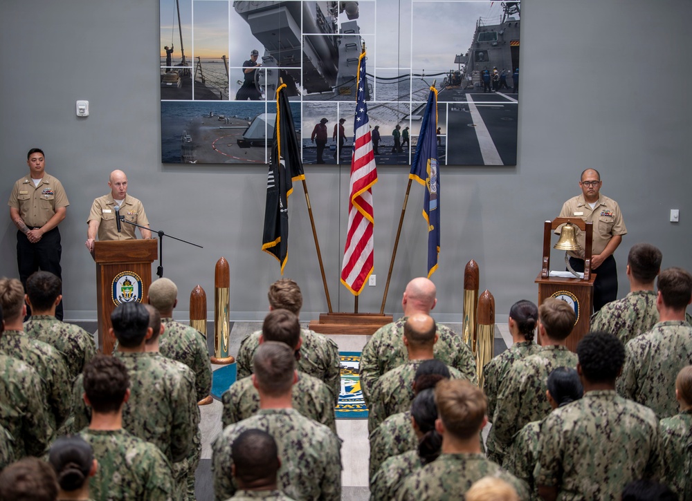 A formal U.S. Navy ceremony is taking place indoors, with a senior officer in khaki service uniform speaking at a podium before a large formation of sailors in Navy Working Uniform (NWU) camouflage. Three flags are displayed prominently — the American flag, a POW/MIA flag, and a Navy flag — alongside decorative shell casings and a ship's bell on a second podium. A large photo collage of naval ship operations serves as the backdrop.