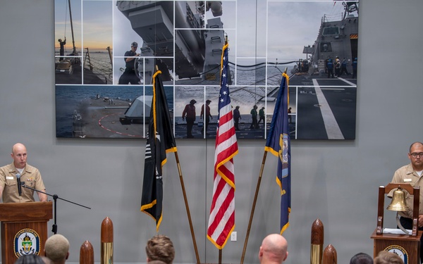A formal U.S. Navy ceremony is taking place indoors, with a senior officer in khaki service uniform speaking at a podium before a large formation of sailors in Navy Working Uniform (NWU) camouflage. Three flags are displayed prominently — the American flag, a POW/MIA flag, and a Navy flag — alongside decorative shell casings and a ship's bell on a second podium. A large photo collage of naval ship operations serves as the backdrop.