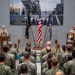 A formal U.S. Navy ceremony is taking place indoors, with a senior officer in khaki service uniform speaking at a podium before a large formation of sailors in Navy Working Uniform (NWU) camouflage. Three flags are displayed prominently — the American flag, a POW/MIA flag, and a Navy flag — alongside decorative shell casings and a ship's bell on a second podium. A large photo collage of naval ship operations serves as the backdrop.