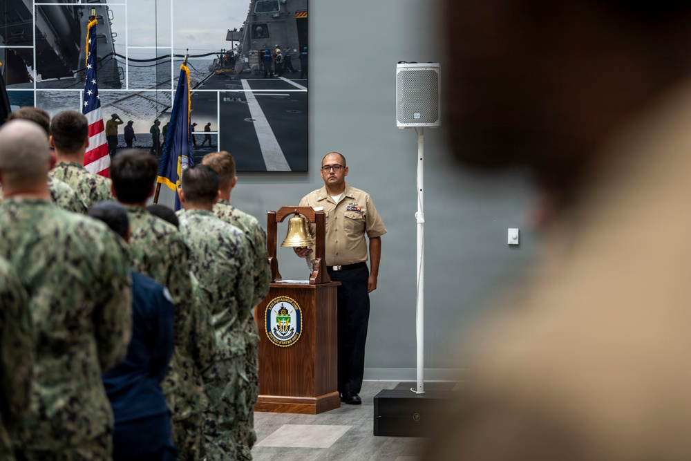 A U.S. Navy ceremonial event is taking place indoors, likely a change of command or retirement ceremony. A sailor in khaki service uniform stands at attention beside a wooden podium bearing a Navy ship's seal and a brass ship's bell. A group of sailors in Navy Working Uniform (NWU) camouflage pattern stand in formation facing the podium, with U.S. and Navy flags visible in the background.