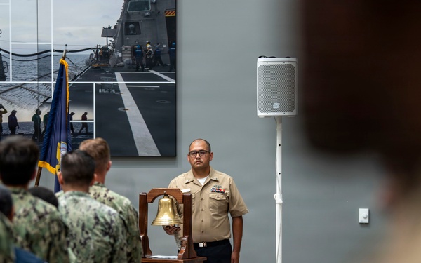 A U.S. Navy ceremonial event is taking place indoors, likely a change of command or retirement ceremony. A sailor in khaki service uniform stands at attention beside a wooden podium bearing a Navy ship's seal and a brass ship's bell. A group of sailors in Navy Working Uniform (NWU) camouflage pattern stand in formation facing the podium, with U.S. and Navy flags visible in the background.