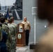 A U.S. Navy ceremonial event is taking place indoors, likely a change of command or retirement ceremony. A sailor in khaki service uniform stands at attention beside a wooden podium bearing a Navy ship's seal and a brass ship's bell. A group of sailors in Navy Working Uniform (NWU) camouflage pattern stand in formation facing the podium, with U.S. and Navy flags visible in the background.