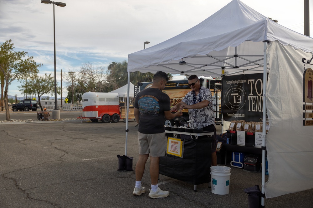 Two men are interacting at an outdoor vendor booth under a white canopy tent, identified as 'Tommy's Tempting Treats' (T3), set up in a parking lot. The vendor appears to be offering a sample or completing a transaction with a customer. The setting suggests an outdoor market or festival event.