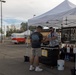 Two men are interacting at an outdoor vendor booth under a white canopy tent, identified as 'Tommy's Tempting Treats' (T3), set up in a parking lot. The vendor appears to be offering a sample or completing a transaction with a customer. The setting suggests an outdoor market or festival event.