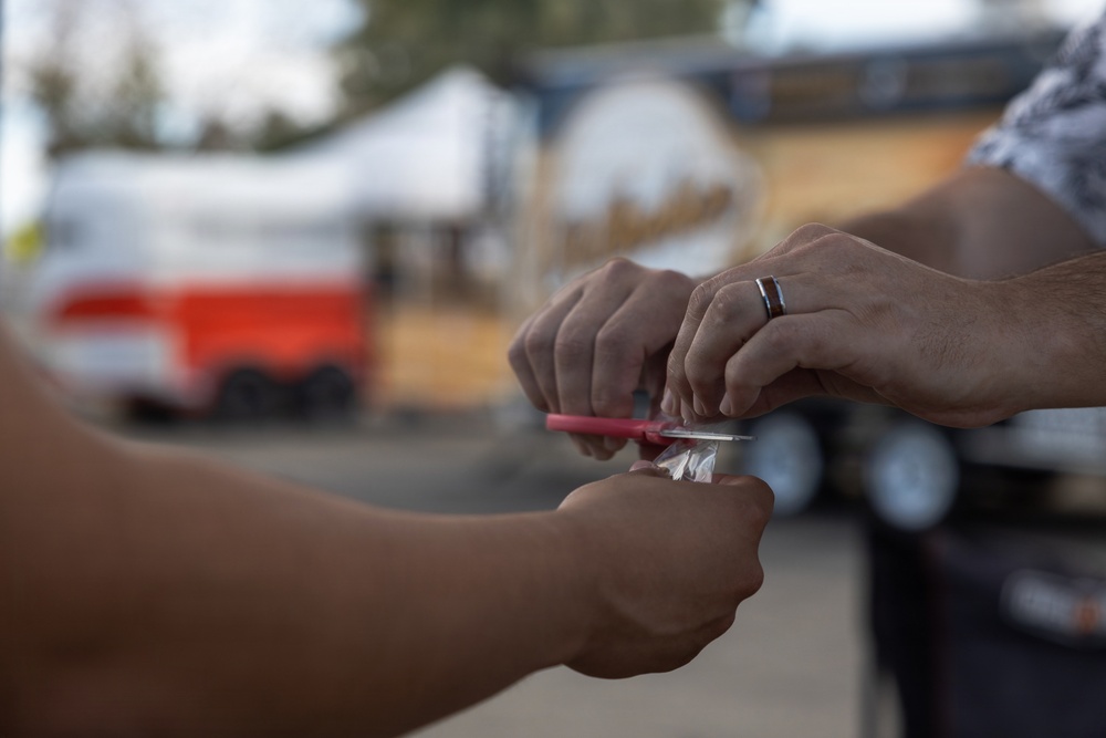 Two individuals are exchanging or handling a small clear plastic bag and a red implement outdoors. One person is wearing a dark ring on their finger. The background shows blurred vehicles and structures in what appears to be a parking or outdoor area.