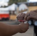 Two individuals are exchanging or handling a small clear plastic bag and a red implement outdoors. One person is wearing a dark ring on their finger. The background shows blurred vehicles and structures in what appears to be a parking or outdoor area.