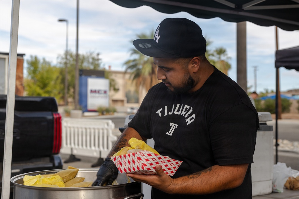 A man wearing a black 'Tijuana' t-shirt and an LA Dodgers cap is serving food at an outdoor food stand or pop-up vendor setup. He is wearing black gloves and handling what appears to be tamales from a large steamer pot, placing them into a red and white checkered food tray. The setting appears to be a parking lot or street-side location with palm trees and commercial buildings visible in the background.