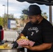 A man wearing a black 'Tijuana' t-shirt and an LA Dodgers cap is serving food at an outdoor food stand or pop-up vendor setup. He is wearing black gloves and handling what appears to be tamales from a large steamer pot, placing them into a red and white checkered food tray. The setting appears to be a parking lot or street-side location with palm trees and commercial buildings visible in the background.