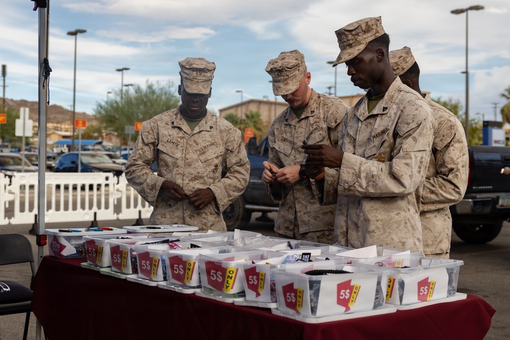 Four U.S. Marines in desert MARPAT camouflage uniforms are browsing items at an outdoor vendor table in what appears to be a parking lot setting. The table is covered with multiple plastic bins labeled with '$5 ONLY' price tags containing various small merchandise items. The setting appears to be a retail parking lot or outdoor market, likely near a military installation.