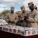 Four U.S. Marines in desert MARPAT camouflage uniforms are browsing items at an outdoor vendor table in what appears to be a parking lot setting. The table is covered with multiple plastic bins labeled with '$5 ONLY' price tags containing various small merchandise items. The setting appears to be a retail parking lot or outdoor market, likely near a military installation.