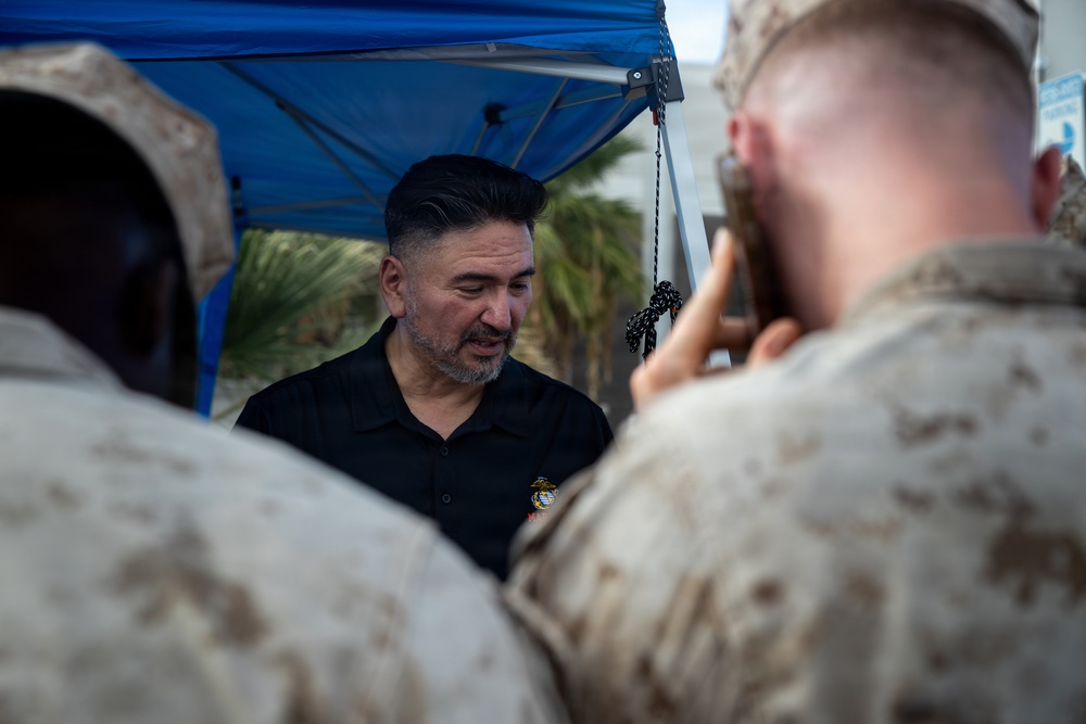 A civilian man wearing a black polo shirt with a Marine Corps emblem is interacting with uniformed U.S. Marines under a blue canopy tent. One Marine in the foreground is photographing or filming the man with a mobile device. The setting appears to be an outdoor military event or meet-and-greet.