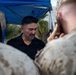 A civilian man wearing a black polo shirt with a Marine Corps emblem is interacting with uniformed U.S. Marines under a blue canopy tent. One Marine in the foreground is photographing or filming the man with a mobile device. The setting appears to be an outdoor military event or meet-and-greet.