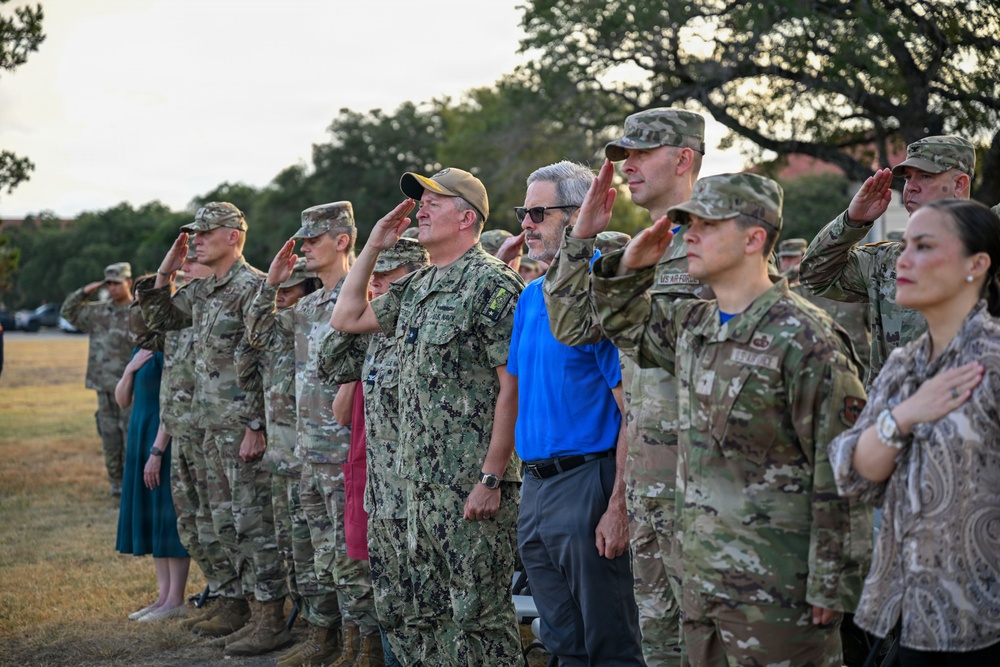 A group of military personnel and civilians stand in formation during what appears to be an outdoor ceremony, likely a flag ceremony or retreat. Most uniformed individuals are rendering a hand salute while two civilians — one in a blue shirt and one in a patterned blouse — stand at attention with hand over heart. Multiple branches of the U.S. military are represented, including Army, Air Force, and Navy.
