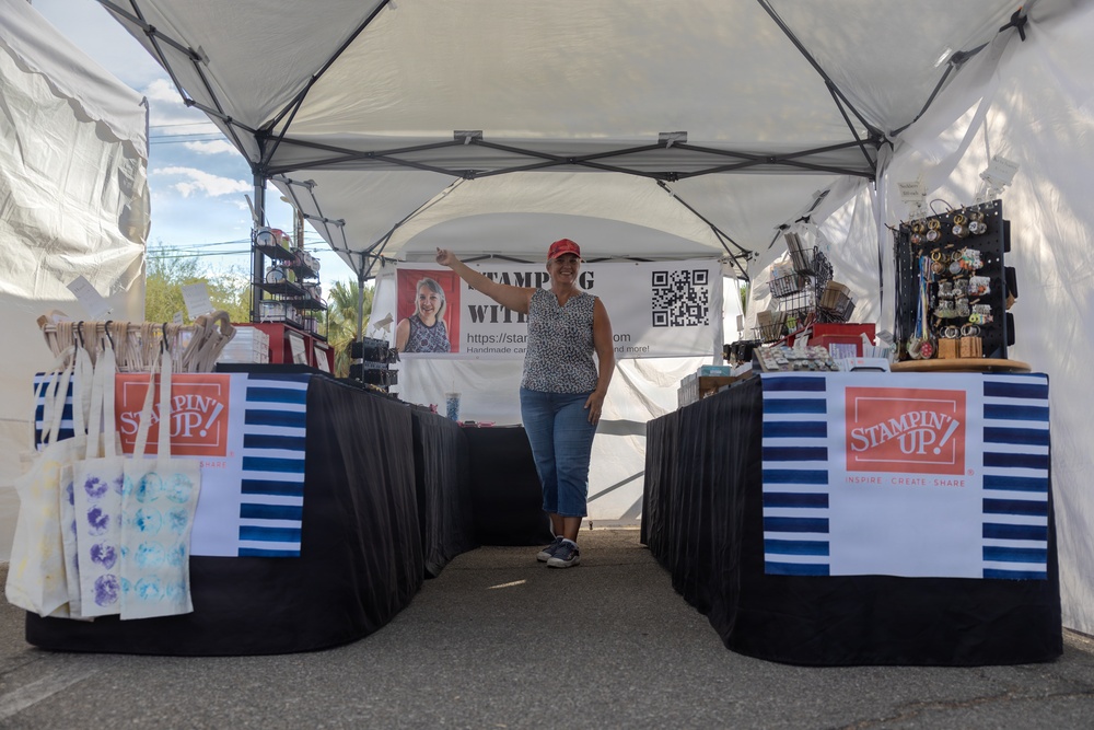 A woman stands inside an outdoor vendor booth at what appears to be a craft fair or market. The booth prominently displays 'Stampin' Up!' branding on both tables, which are covered with black tablecloths and navy/white striped panels. The booth features handmade cards, tote bags, jewelry, and other craft items for sale.