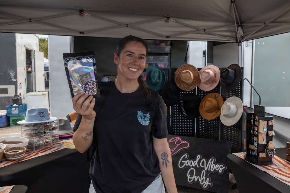 A smiling woman stands at an outdoor market vendor booth under a gray canopy tent, holding up a holographic mylar pouch product toward the camera. The booth displays an assortment of felt hats, jewelry, and accessories against a black pegboard backdrop. A neon sign reading 'Good Vibes Only' is prominently visible on the table.