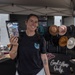 A smiling woman stands at an outdoor market vendor booth under a gray canopy tent, holding up a holographic mylar pouch product toward the camera. The booth displays an assortment of felt hats, jewelry, and accessories against a black pegboard backdrop. A neon sign reading 'Good Vibes Only' is prominently visible on the table.
