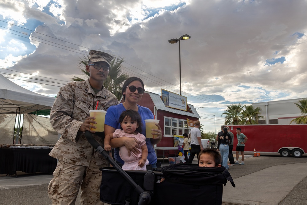 A U.S. Marine in desert camouflage uniform poses with his family at what appears to be an outdoor community or base event. The group includes a woman holding a baby and a young child sitting in a stroller, with both adults holding cold drinks. The setting features food vendor stalls, palm trees, and a partly cloudy sky in what appears to be a desert region.
