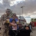 A U.S. Marine in desert camouflage uniform poses with his family at what appears to be an outdoor community or base event. The group includes a woman holding a baby and a young child sitting in a stroller, with both adults holding cold drinks. The setting features food vendor stalls, palm trees, and a partly cloudy sky in what appears to be a desert region.