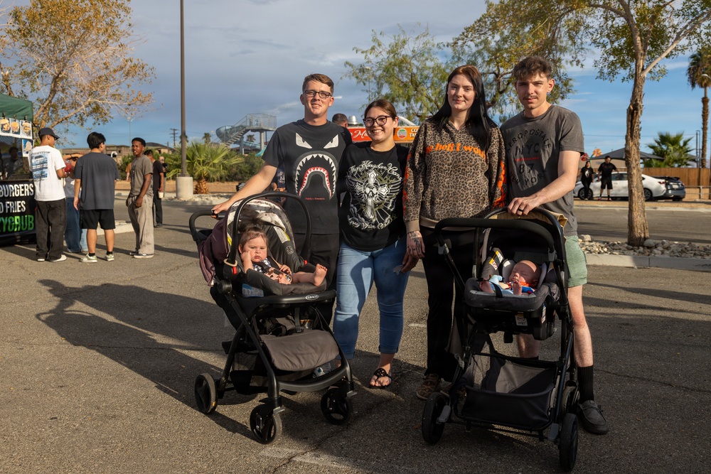 Four young adults pose together at an outdoor event, each couple accompanied by an infant in a Graco car seat stroller. The group is standing in a parking lot area with food trucks and other attendees visible in the background. The setting appears to be a casual community or street fair event during late afternoon.