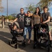 Four young adults pose together at an outdoor event, each couple accompanied by an infant in a Graco car seat stroller. The group is standing in a parking lot area with food trucks and other attendees visible in the background. The setting appears to be a casual community or street fair event during late afternoon.