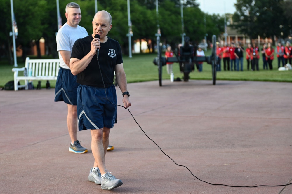 A bald man in athletic wear is speaking into a wired microphone at what appears to be an outdoor event on a military or institutional campus. He is wearing a black t-shirt with a military insignia and navy blue shorts. In the background, a group of people dressed in red shirts are gathered near flagpoles and a cannon monument.