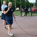 A bald man in athletic wear is speaking into a wired microphone at what appears to be an outdoor event on a military or institutional campus. He is wearing a black t-shirt with a military insignia and navy blue shorts. In the background, a group of people dressed in red shirts are gathered near flagpoles and a cannon monument.