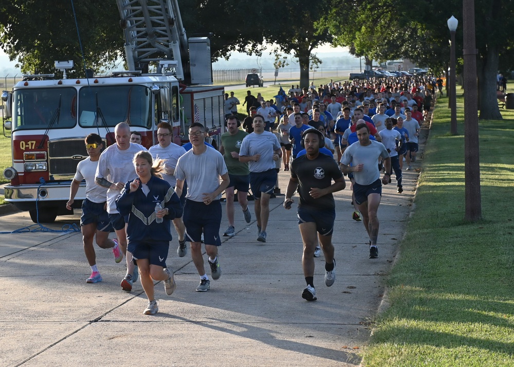 A large group of military personnel, likely U.S. Air Force members based on visible insignia and PT gear, are participating in a group run along a paved path on what appears to be a military installation. A fire truck labeled 'Q-17' is parked alongside the route, possibly as part of a memorial or ceremonial run. The event appears to involve hundreds of participants stretching far into the background.