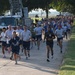 A large group of military personnel, likely U.S. Air Force members based on visible insignia and PT gear, are participating in a group run along a paved path on what appears to be a military installation. A fire truck labeled 'Q-17' is parked alongside the route, possibly as part of a memorial or ceremonial run. The event appears to involve hundreds of participants stretching far into the background.