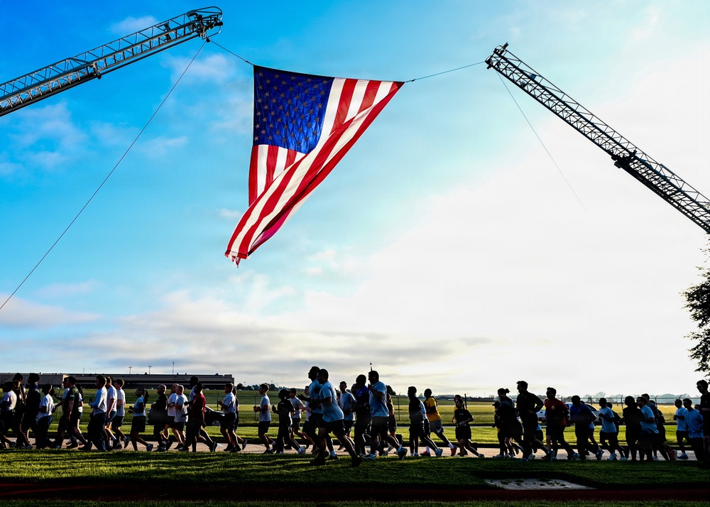 A large group of people are running or walking beneath an oversized American flag suspended between two fire truck aerial ladders. The event appears to be a commemorative or patriotic run, likely a memorial or community fitness event. The image is taken at ground level during early morning or late afternoon light, creating dramatic silhouettes against a blue sky.