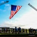 A large group of people are running or walking beneath an oversized American flag suspended between two fire truck aerial ladders. The event appears to be a commemorative or patriotic run, likely a memorial or community fitness event. The image is taken at ground level during early morning or late afternoon light, creating dramatic silhouettes against a blue sky.