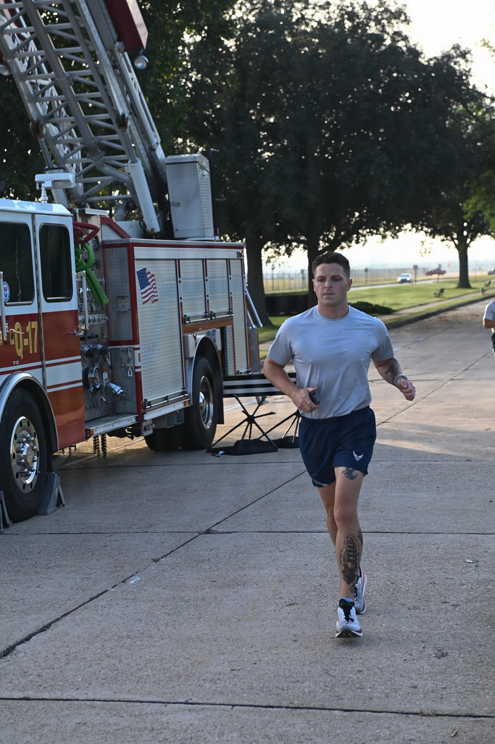 A man in athletic wear is running on a concrete path alongside a red fire truck labeled 'Q-17' with an American flag decal. The setting appears to be a military base or airfield, suggested by the open grounds, trees, and the U.S. Air Force logo visible on the runner's shorts. The runner has visible tattoos on his arm and leg and appears to be participating in a fitness run or memorial event.