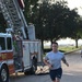 A man in athletic wear is running on a concrete path alongside a red fire truck labeled 'Q-17' with an American flag decal. The setting appears to be a military base or airfield, suggested by the open grounds, trees, and the U.S. Air Force logo visible on the runner's shorts. The runner has visible tattoos on his arm and leg and appears to be participating in a fitness run or memorial event.