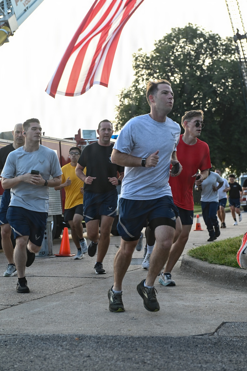 A group of runners participates in an organized run event, likely a military fitness run or memorial run based on the U.S. Air Force insignia visible on several participants' shirts and shorts. An American flag is prominently displayed overhead, suspended from what appears to be a fire truck ladder. Orange traffic cones mark the course route on a paved road.